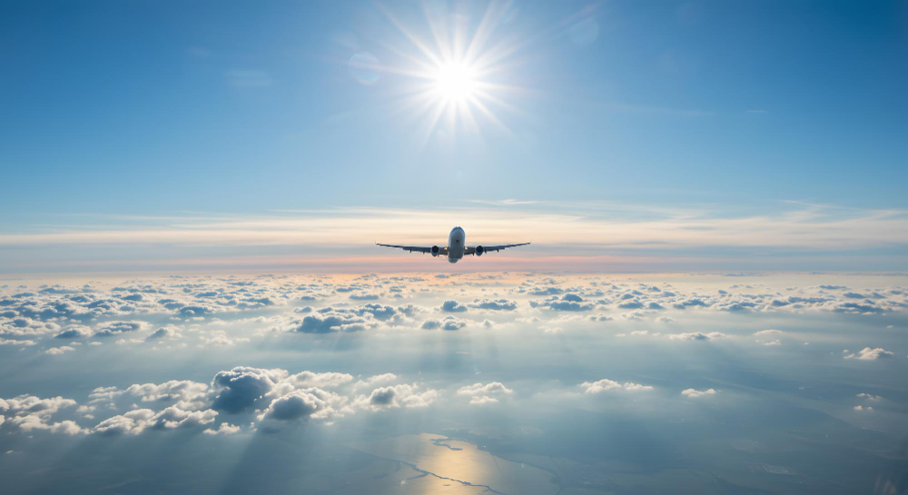 Um avião por cima das nuvens em um dia de sol. A imagem representa apps para encontrar passagens aéreas baratas.